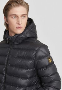 Giacca a vento nera con una superficie lucida, trapuntatura orizzontale, cappuccio e una toppa del logo sulla manica sinistra. Chiusura frontale con zip.