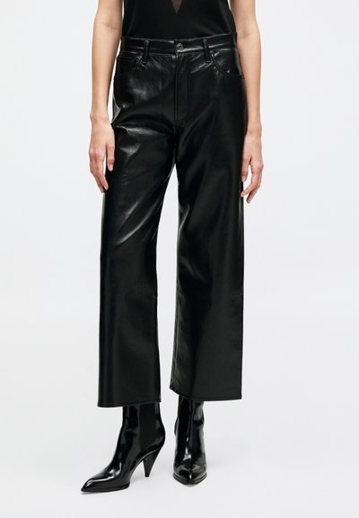 Donna che indossa pantaloni neri a gamba larga in ecopelle abbinati a stivaletti neri con punta appuntita e top nero senza maniche.