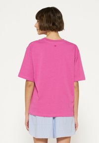 T-shirt di cotone rosa con maniche corte e collo a giro. Presenta un piccolo dettaglio a linea orizzontale sul retro. Indossato con pantaloni corti a righe blu.