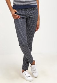 Person som bär marinblå mönstrade slim-fit-byxor och vita sneakers med kardborreband, står med benen korsade och en hand i fickan.