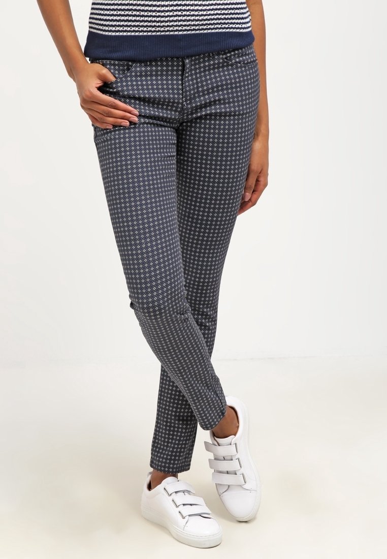 Person som bär marinblå mönstrade slim-fit-byxor och vita sneakers med kardborreband, står med benen korsade och en hand i fickan.