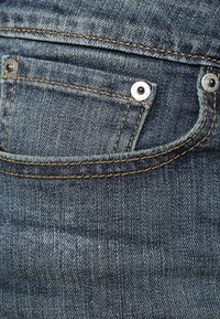 Denim-kangasta syvässä sinisessä värissä, jossa on takatasku metallinitteillä ja kontrastiväriset vaaleanruskeat ompeleet reunan ympäri.