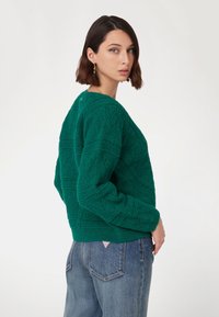 Maglione a maglia verde con texture e motivo geometrico, caratterizzato da un ampio scollo, spalle scese e polsini a costine, indossato con jeans blu.