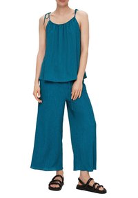 Türkise ärmellose Bluse mit verstellbaren Trägern, kombiniert mit lockeren, weit geschnittenen Hosen. Leichter Stoff mit subtiler Textur. Schwarze Sandalen runden den Look ab.