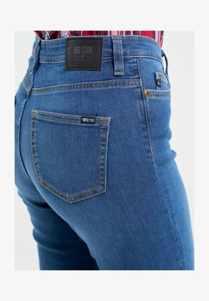 Tumši zili džinsi ar augsto jostu, ar dzelteno šuvju un melno ādas zīmolu uz aizmugures. Iekļauti divi aizmugurējie kabatas.