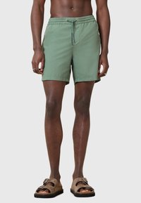 Pantaloni da bagno leggeri di colore verde con cintura elastica e cordoncino. Indossati con sandali marroni senza strap con una doppia chiusura a fibbia.