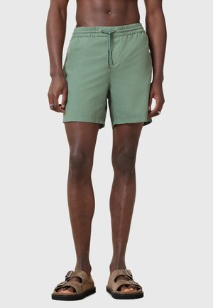 Gröna lätta simshorts med elastisk midja och dragsko. Bärs med bruna slip-on sandaler med en design av dubbel spänne.