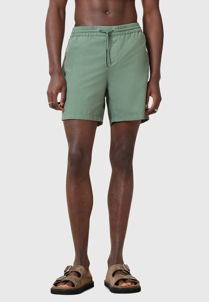 Pantaloni da bagno leggeri di colore verde con cintura elastica e cordoncino. Indossati con sandali marroni senza strap con una doppia chiusura a fibbia.