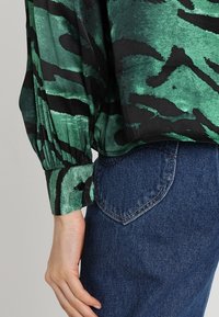 Green and black patterned long-sleeve blouse with textured fabric and buttoned cuffs, paired with blue denim jeans featuring a back pocket.