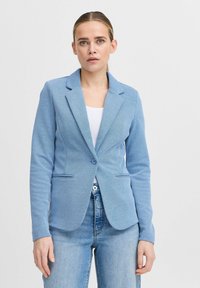 Světle modrý blazer vyrobený z texturované látky, se zapínáním na jeden knoflík, s klopami a dvěma předními kapsami. Nositý přes bílou halenku a džínové kalhoty.