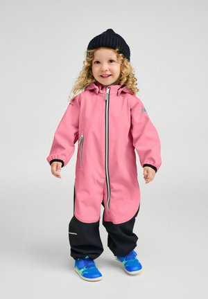 Małe dziecko z kręconymi włosami ubrane w różowo-czarny wodoodporny kombinezon, czapkę z dzianiny w kolorze czarnym oraz niebieskie trampki, stojące i uśmiechające się.