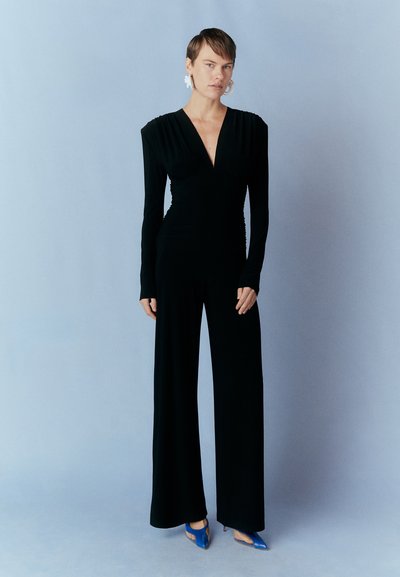 Black long-sleeve jumpsuit with deep V-neck, gathered sides, and wide-leg silhouette, paired with blue heels, set against a light blue background.