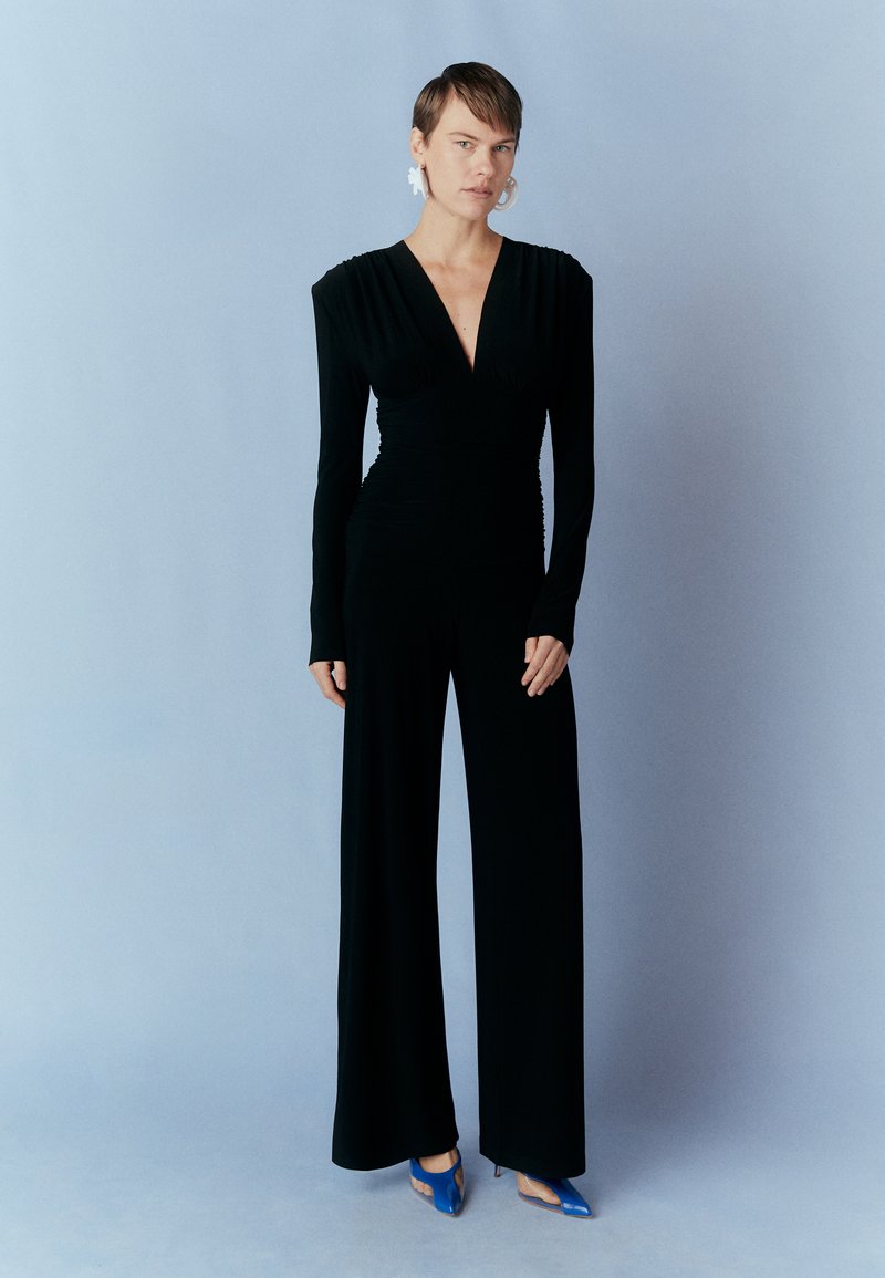 Zwart langelijner jumpsuit met diepe V-hals, gathered zijkanten en een brede broekpasvorm, gecombineerd met blauwe hakken, tegen een lichtblauwe achtergrond.