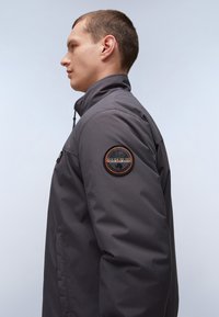 Grå midilængde jakke med høj krave, lynlås lukning, sidelommer og et broderet logo patch på venstre ærme. Glat tekstur.