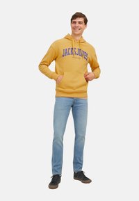 Gelbes Kapuzensweatshirt mit blauer "JACK & JONES"-Druckschrift, Kängurutasche, gerippte Bündchen, kombiniert mit hellblauen Jeans und schwarzen Turnschuhen.