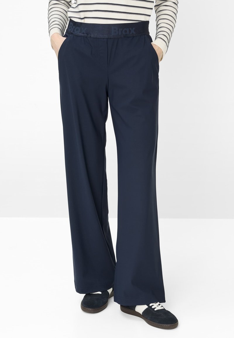 Persoon draagt marineblauwe broek met wijde pijpen en elastische tailleband met het label "Brax", gestreept shirt met lange mouwen en zwart-witte sneakers.