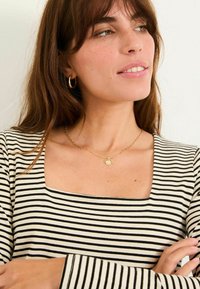 Gestreiftes Langarmoberteil in Schwarz und Weiß mit quadratischem Ausschnitt. Eine goldene Kettenhalskette mit einem runden Anhänger wird getragen.