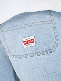 Lys blå denimjeans baklomme med hvit etikett med teksten «DENIM ONLY & SONS ARTISANS» i svart og rødt mot en hvit overflate.