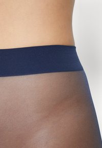 Námorně modrý pas vedoucí k průhledným hnědým punčochám, které mají měkkou texturu a odolný pletený design. Přechod od neprůhledného k průhlednému.