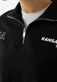 Pullover in fleece nero con colletto zip, caratterizzato da testo bianco stampato "KANSAS" e un piccolo logo. Texture liscia e design sportivo.
