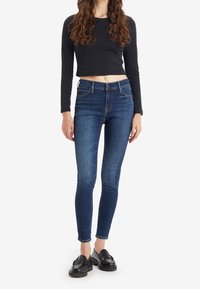 Mujer vistiendo jeans skinny azul oscuro, top negro de manga larga recortada y mocasines negros, de pie contra un fondo blanco liso.