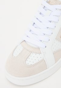 Vita läder och beige mocka sneakers med perforerad detaljer, vita snören och en texturerad gummisula. Varumärkeslogotyp i guld på sidan.