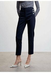 Pantalons tailleur bleu marine à taille haute, avec poches latérales et boutons décoratifs. Associés à des escarpins blancs à lanières avec un détail en nœud.