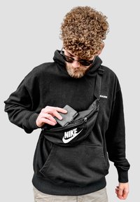 Black Nike fanny pack worn crossbody; fabric material with smooth texture; features zippered pockets and logo, paired with a black hoodie.