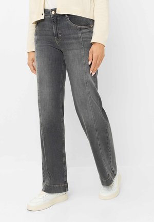 Persoon draagt donkergrijze rechte jeans, een crème cardigan, witte sneakers, staand tegen een effen lichte achtergrond.