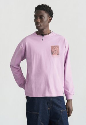 Fiatal férfi, rózsaszín hosszú ujjú pólóban, amelyen grafikai mintázat látható a mellkasán, valamint sötétkék farmert visel, egyik kezét a háta mögött tartva áll.