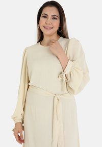 Cream long-sleeve dress with gathered sleeves and a tied belt at the waist. Lightweight fabric with a soft texture. Simple neckline design.
