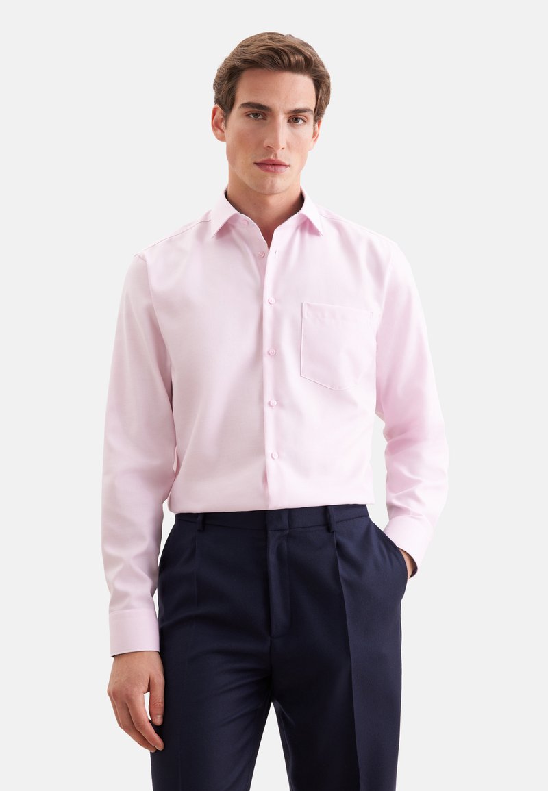 Jeune homme portant une chemise rose clair à manches longues avec poche poitrine et pantalon bleu marine foncé, debout avec une main dans la poche.