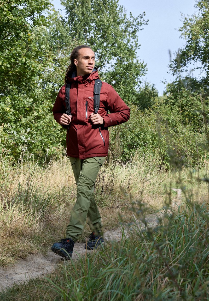 Chaqueta impermeable roja con bolsillos, pantalones verdes y botas de senderismo negras. Caminando por un camino de tierra rodeado de hierba alta y arbustos.