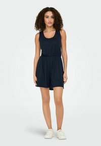 Combinaison sans manches bleu marine avec taille élastique, tissu doux et coupe décontractée. Associée à des baskets blanches, se tenant devant un fond neutre.