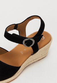 Sandale à talon compensé en suède noire avec bride de cheville et boucle métallique. Talon compensé en jute, semelle intérieure lisse en brun avec logo embossé.