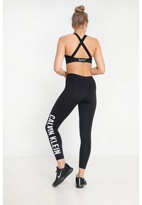 Czarny stanik sportowy z krzyżującym się tyłem od Nike, w połączeniu z czarnymi legginsami z dużym białym napisem "CALVIN KLEIN" oraz czarnymi butami sportowymi.