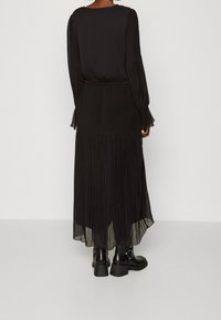 Svart plisserad maxiklänning med långa ärmar, figurnära midja, flödande kjol och volangkantade ärmslut. Bärs med svarta ankelboots.
