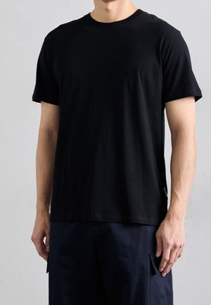 Man draagt een eenvoudig zwart T-shirt met korte mouwen en een ronde hals, en donkere broek, staand tegen een lichtgrijze achtergrond.