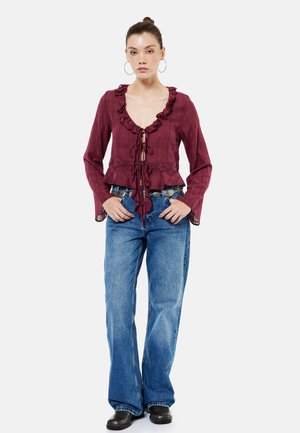 Ung kvinde iført en vinrød bluse med flæser og bindelukninger, brede blå jeans, sorte sko og store øreringe i ringform, stående med hænderne i lommerne.