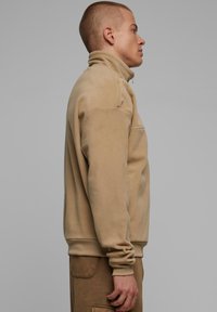 Beige fleecejakke med høy krage og glidelås, med avslappet passform og ribbestrikkede mansjetter, sett i sideprofil.