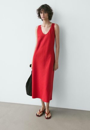 Frau mit lockigem Haar, die ein ärmelloses rotes Midikleid und rote Flip-Flops trägt, eine schwarze Tasche in der Hand haltend, vor einer weißen Wand stehend.