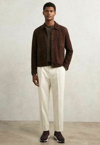 Pruun nahkjakker nööpide sulgemisega, kahe eesmise taskuga, kantud tumeda pruuni särgi peal. Valged kohandatud püksid ja tume spordijalatsid täiendavad riietust.