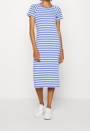 Frau trägt ein blau-weiß gestreiftes Midikleid mit kurzen Ärmeln und weißen Sneakers, steht vor einem schlichten Hintergrund.