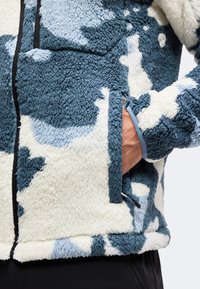 Personne portant une veste polaire à motifs bleu et blanc, la main dans la poche latérale et une fermeture éclair noire visible.