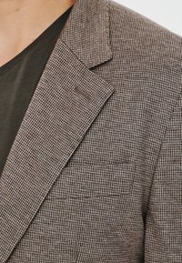 Brązowy blezer w wzór houndstooth z wyciętymi klapami, kieszenią na piersi i teksturowanym materiałem, zestawiony z ciemnozieloną koszulką pod spodem.