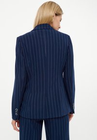 Blazer de rayas azul marino con cuello de muesca, corte entallado, abertura trasera simple y puños con tres botones y acentos metálicos.