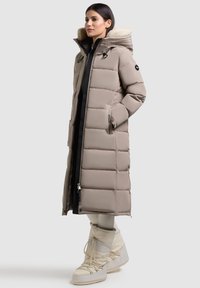 Geacă puffer lungă, bej, cu glugă căptușită cu blană, model cu matlasare diagonală, buzunare laterale și închidere cu fermoar. Asortată cu cizme de culoare crem.