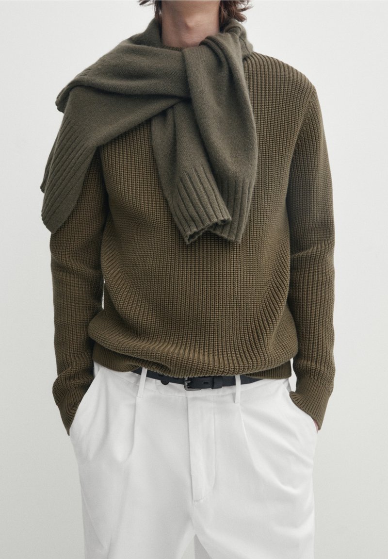 Maglione lavorato a maglia verde oliva con texture a coste, abbinato a una sciarpa dello stesso colore. I pantaloni bianchi e la cintura nera completano l'outfit.