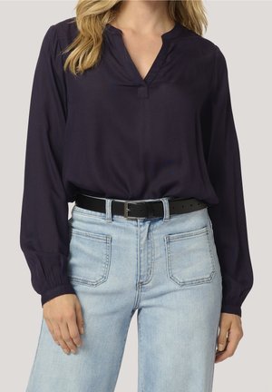 Femme portant un chemisier bleu marine foncé à manches longues, rentré dans un jean taille haute bleu clair avec une ceinture noire.