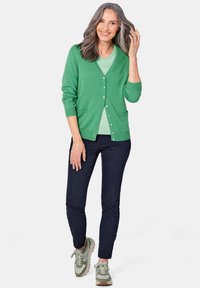 Grüne Strickjacke mit Knöpfen, leichtes Material, zwei vordere Taschen, getragen über einem hellgrünen Oberteil und kombiniert mit dunklen Jeans und Sneakers.